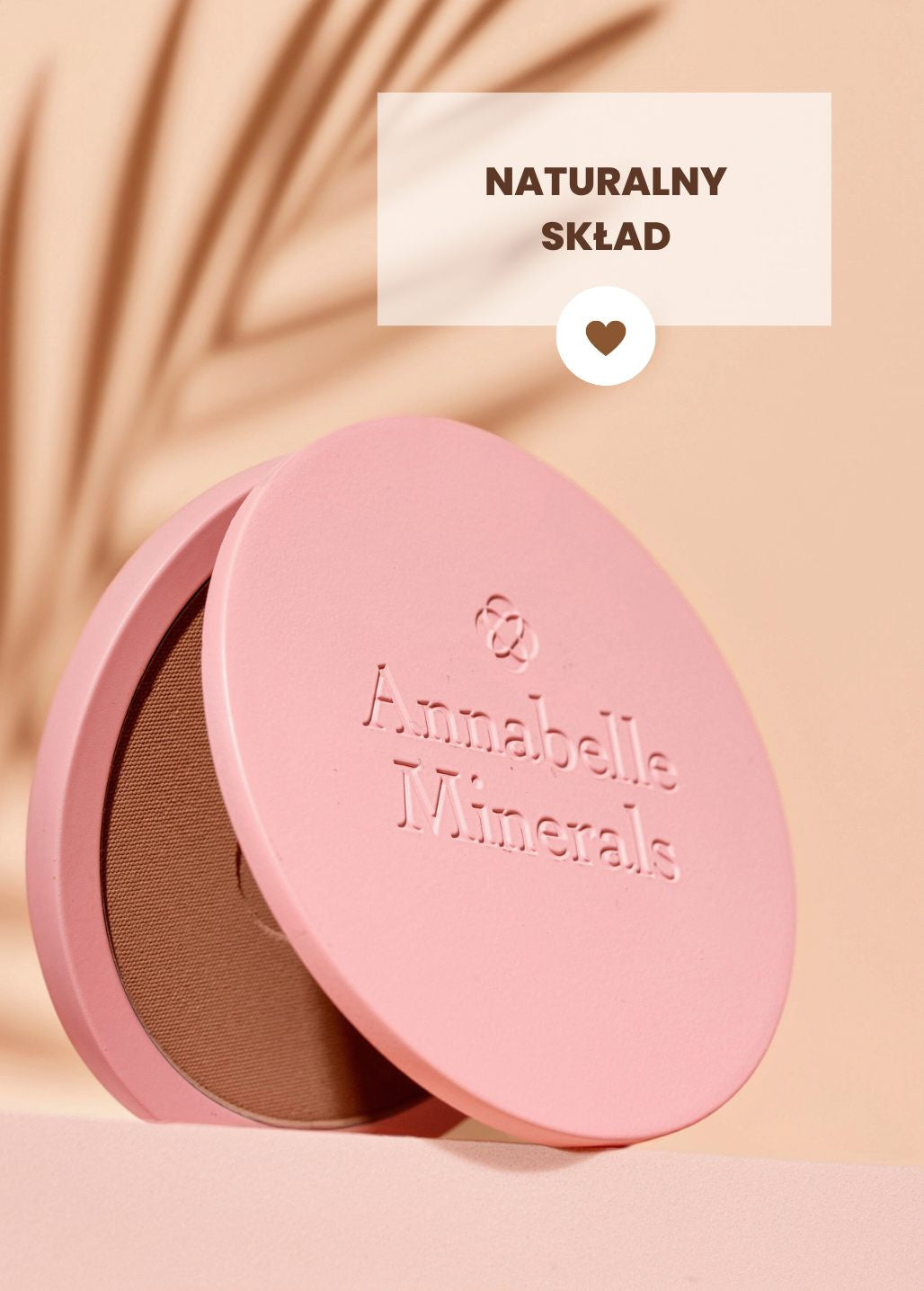 Różowy kompakt z bronzerem prasowanym Annabelle Minerals z etykietą naturalny skład na tle w ciepłych odcieniach