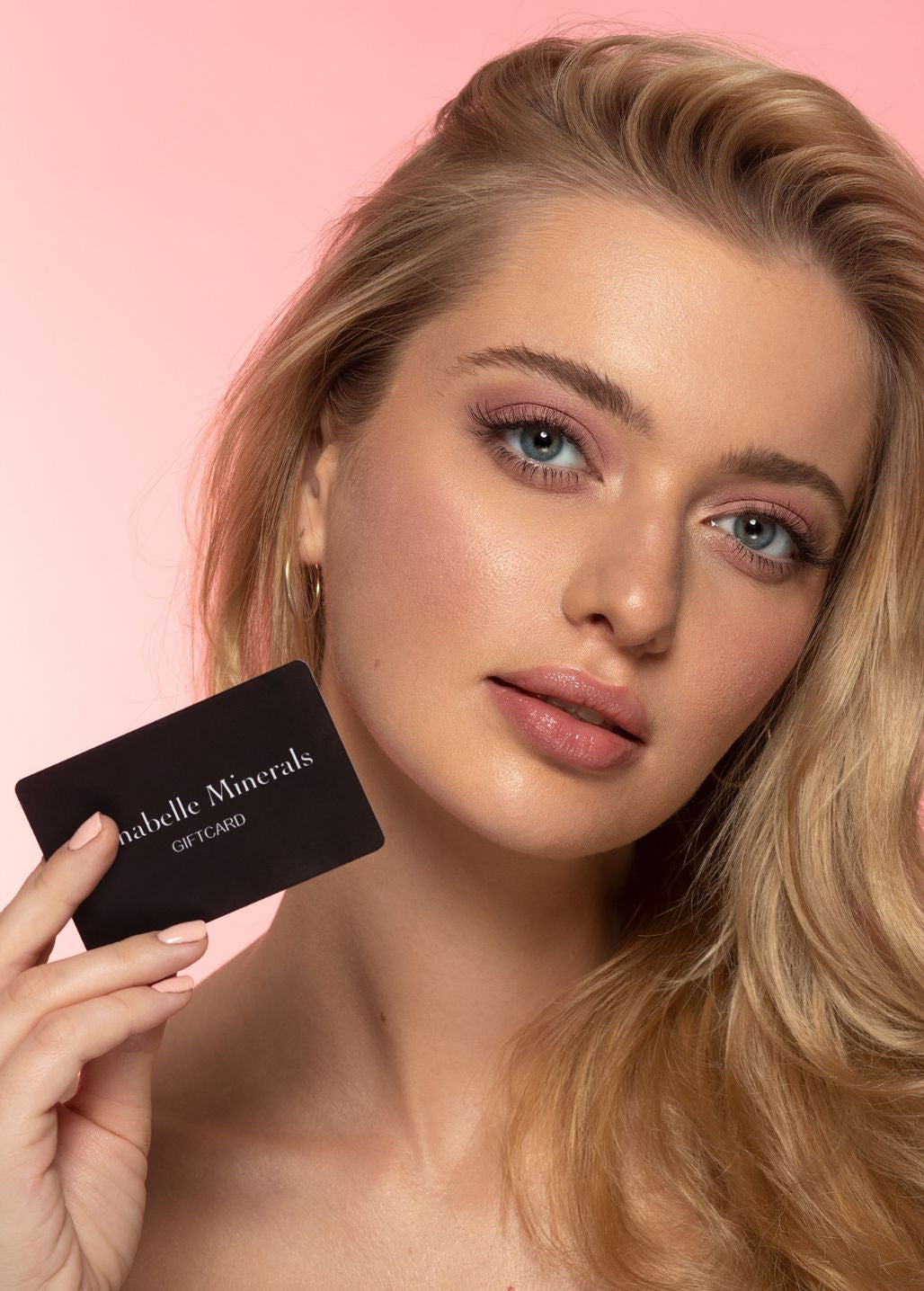 En person med långt, blont hår håller i ett svart presentkort från Annabelle Minerals mot en rosa bakgrund