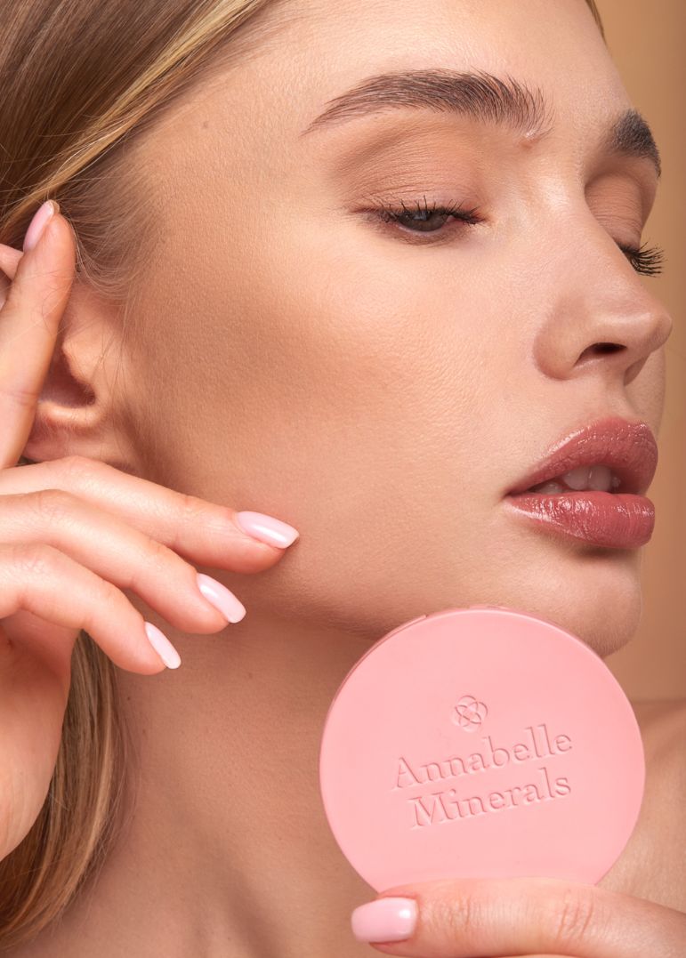 Profil osoby z subtelnym makijażem trzymającej różowy produkt Annabelle Minerals na tle w pastelowych kolorach