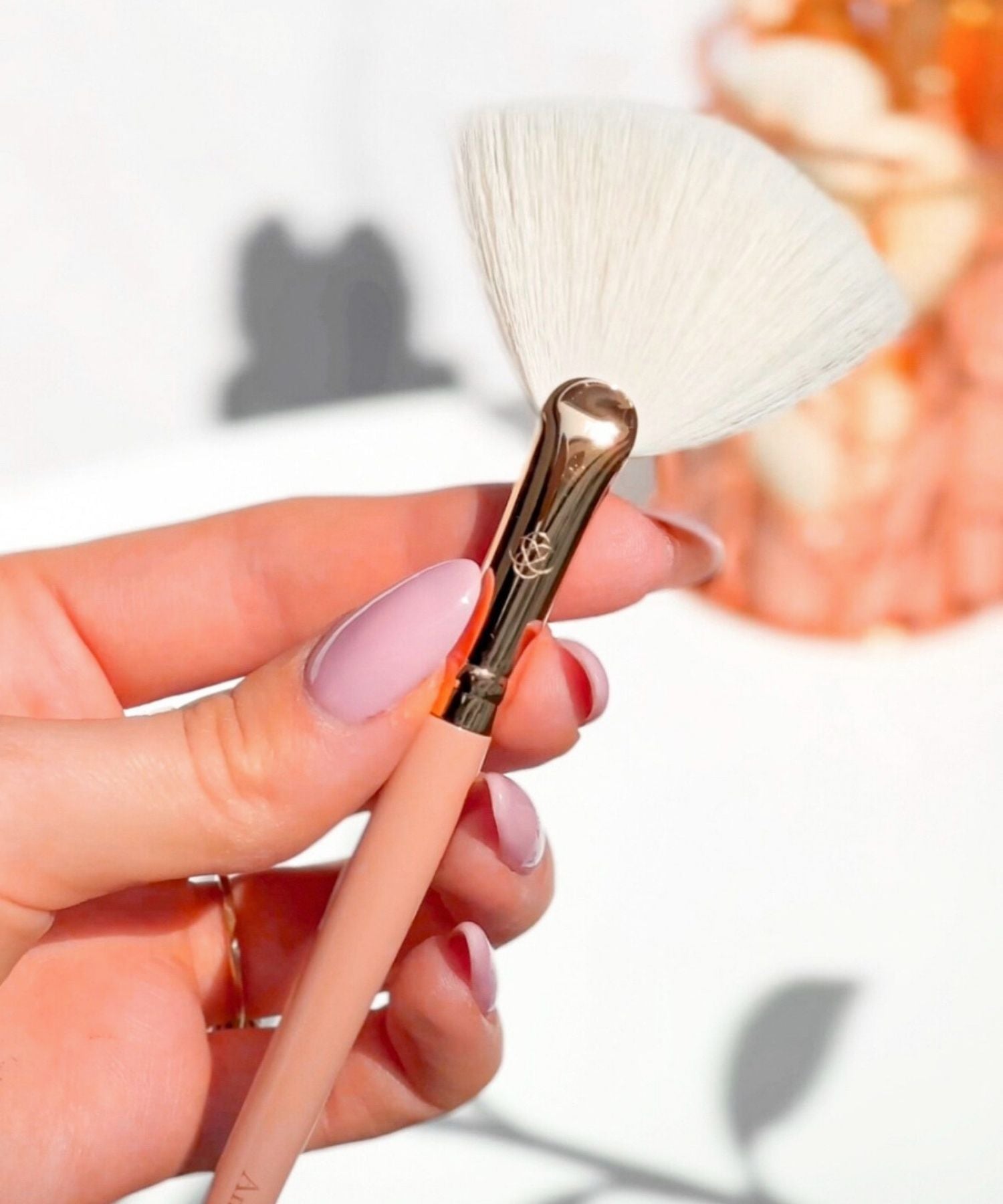 Pędzel wachlarz do nakładania rozświetlacza Annabelle Minerals z rozłożystym włosiem i długą, różową rączką, trzymany w dłoni z jasnymi, pastelowymi paznokciami