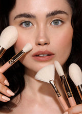Osoba z jasną skórą i ciemnymi włosami trzyma miękkie pędzle do makijażu Annabelle Minerals, podkreślając naturalny look.